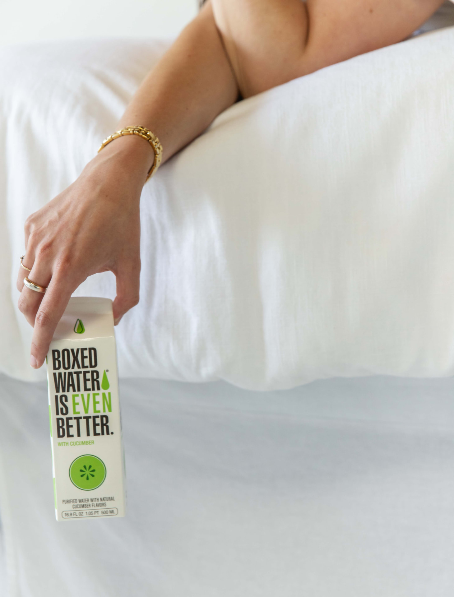 Cropped frame of a hand resting a cucumber Boxed Water carton on white bedding