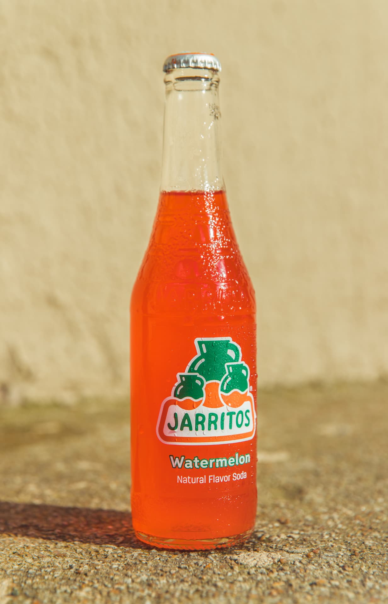 Watermelon Jarritos bottle standing in daylight against a warm wall