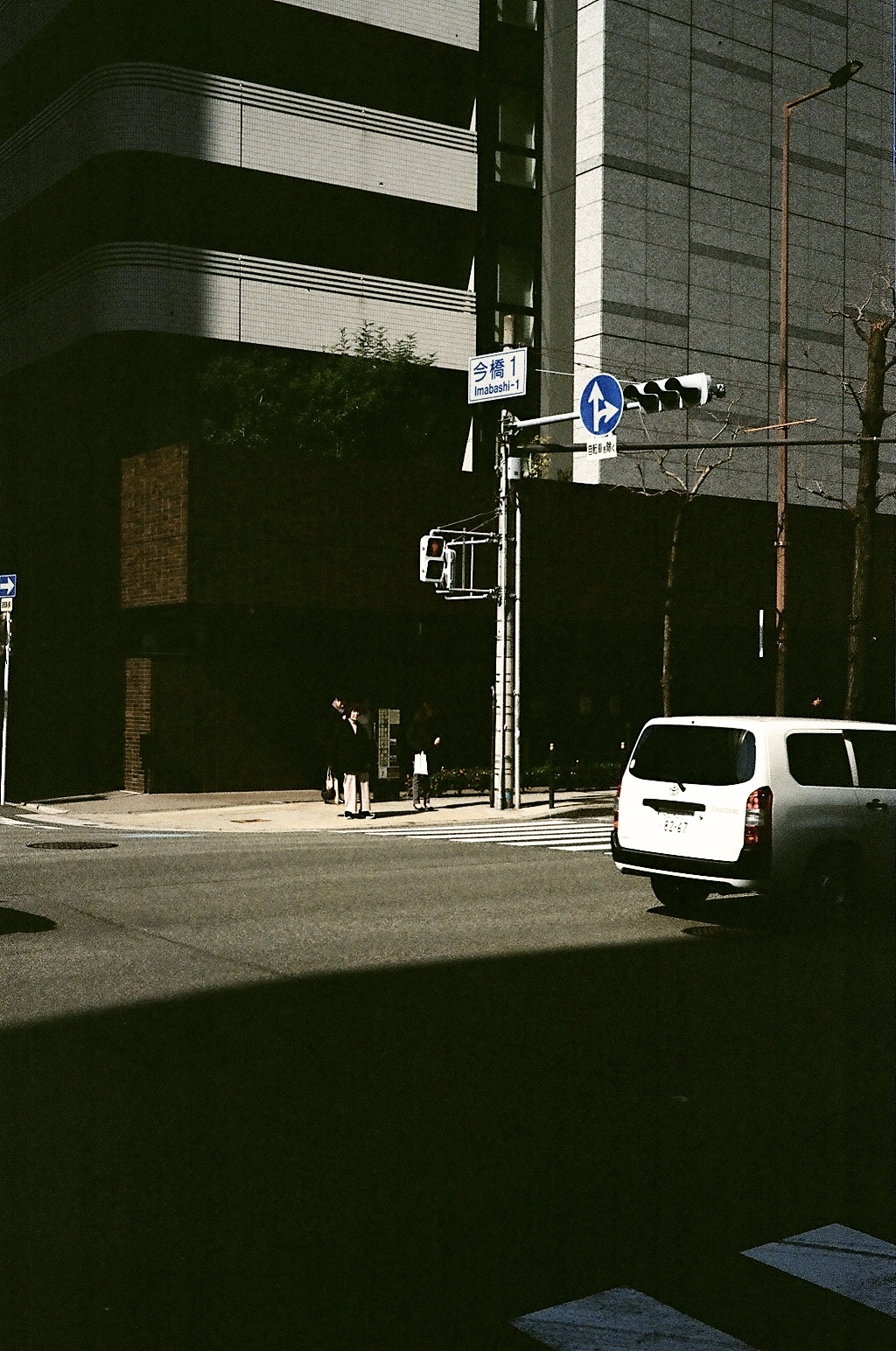 Figures waiting at a sunlit crossing beside a shadowed office block