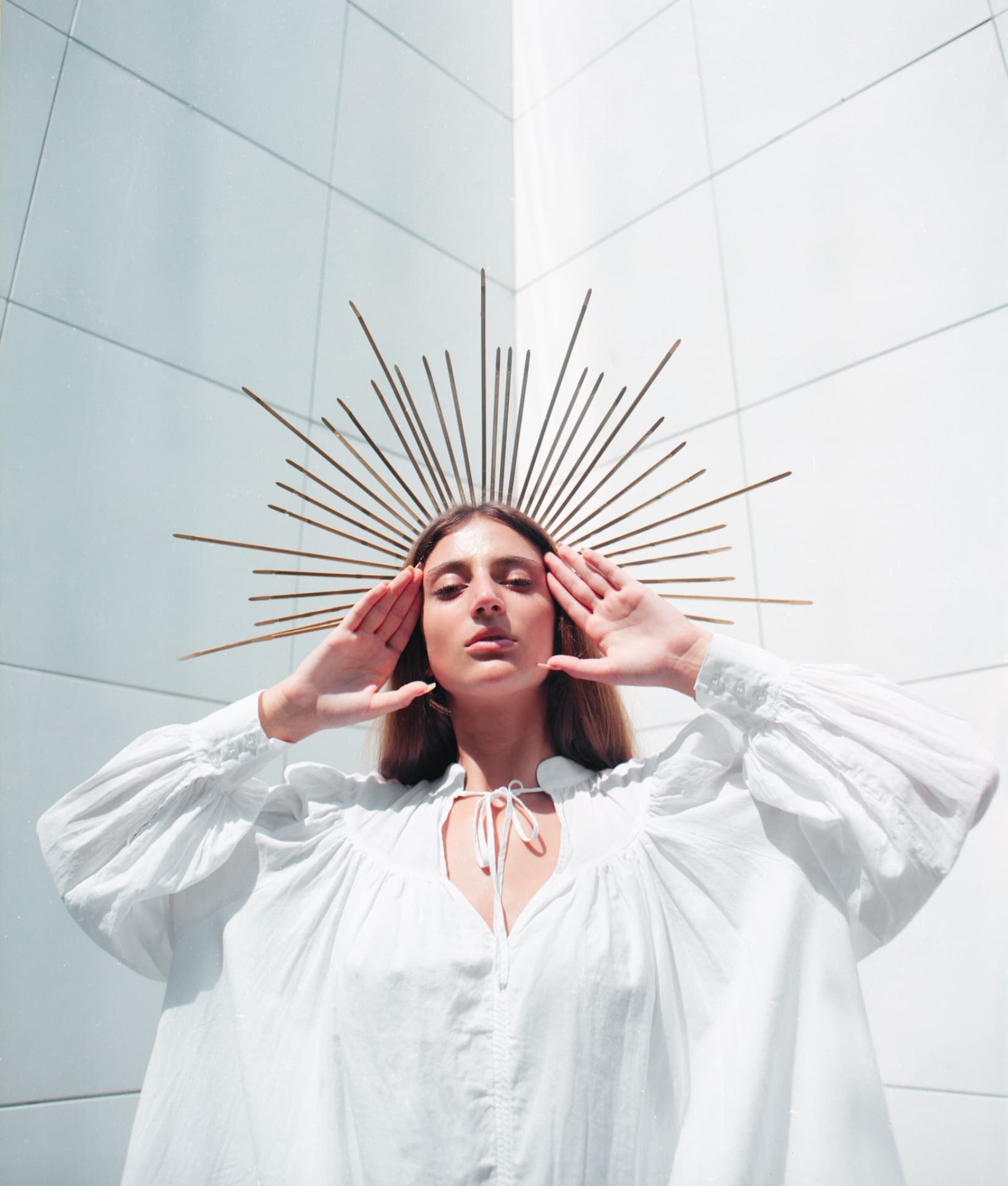 Portrait in a white smock with a brass sunburst halo against a pale wall