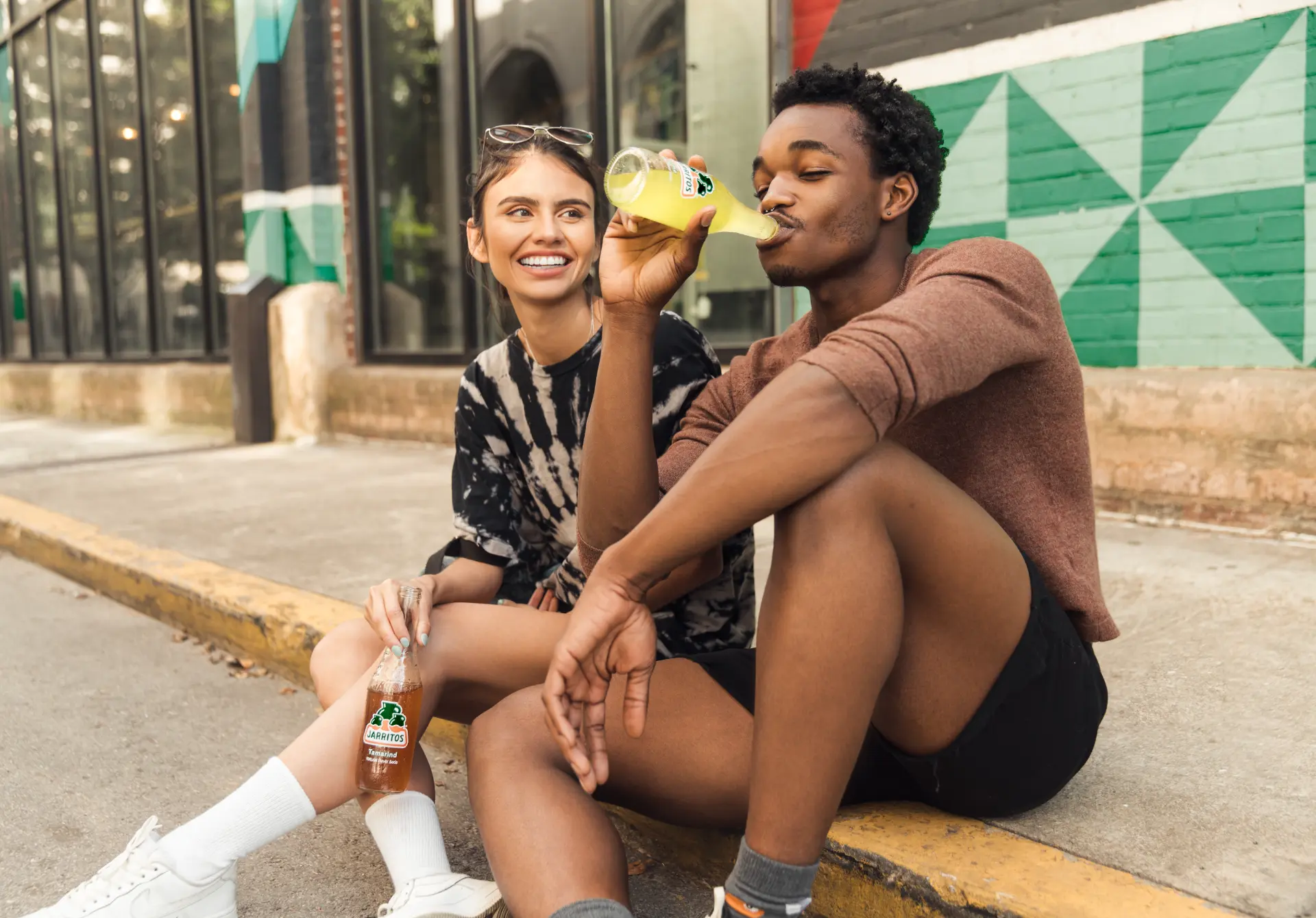 Two people sitting on a curb drinking Jarritos in late afternoon light